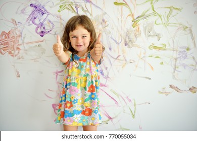 Curly cute little toddler girl finished painting with paints color and brush on the wall. Works of child. Thumbs up. - Powered by Shutterstock