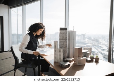 Curly female engineer working on building complex prototype project of residential or business district, measuring blueprint city model using compass at office with panoramic windows - Powered by Shutterstock