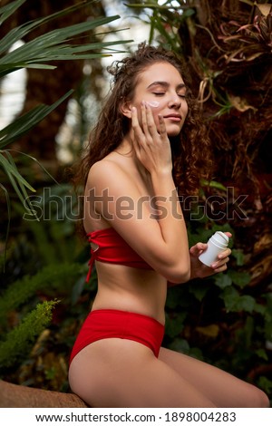 curly female in red bikini applying cream on face in the forest,, enjoys soft skin using organic cosmetics. beauty concept