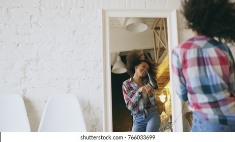 Curly Funny African American Girl Dancing And Singing With Hair Dryer In Front Of Mirror At Home