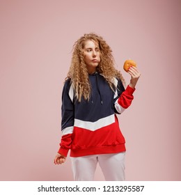 Curly Girl With Blonde Long Hair In Full Height On Pink Background. One Hand Holding Burger, Chewing, Greedily Looking At Him. Concept Of Fast Food