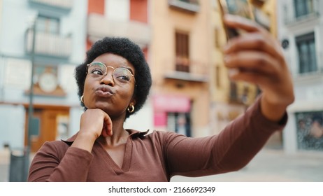 Curly Girl Wearing Glasses  Sending Kiss Taking Selfie On The Street. Cute African Woman Making Content For Social Media Outdoors