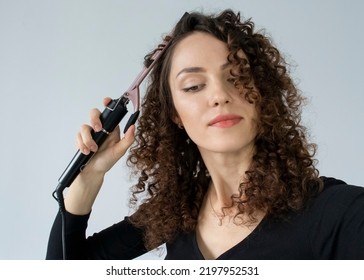 Curly hair. Beautiful smiling woman with curly hair, curls hair, uses curling iron tongs to get fine curls. Hair style and hairdressing tools.