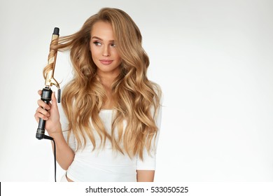 Curly Hair. Beautiful Smiling Woman With Long Blonde Wavy Hair Ironing It, Using Curling Iron. Happy Girl With Gorgeous Healthy Smooth Hair Using Curler For Perfect Curls. Hairstyle And Hairdressing