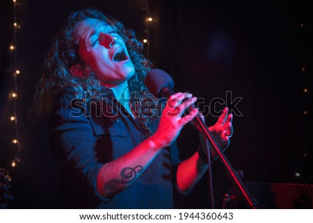 Curly hair woman performing music show with microphone. Rock pop female star singer performer with treble clef tattoo on her arm. Lights music concert