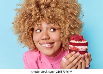 Curly Haired Beautiful Woman Bites Lips Looks At Appetizing Piece Of Cake With Raspberries Has Sweet Tooth Feels Temptation To Eat Delicious Dessert Isolated Over Blue Background. Sugar Addiction