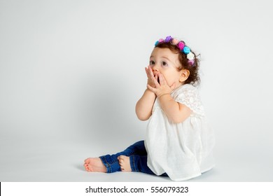 A curly haired, brunette baby wearing a colorful, floral headband sits on a white background with hands to her mouth.  She is blowing kisses but looks surprised.  