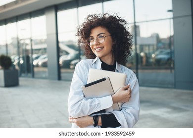 Curly Haired Business Lady Is Embracing And Holding A Laptop While Posing With Glasses In Front Of A Building