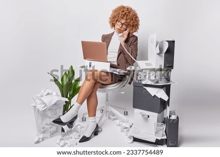 Curly haired elegant businesswoman engages in phone conversation via stationary phone works on laptop computer surrounded by chaotic mess of crumpled papers wears formal clothes poses in office
