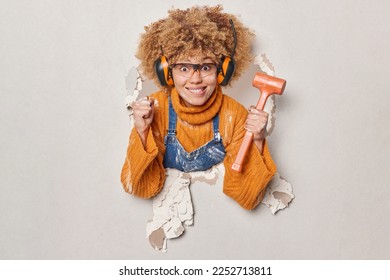 Curly haired female builder holds hammer clenches fist maintainces house renovation service wears glasses headphones on ears dressed in orange jumper and overalls breaks through plaster wall