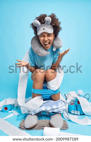 Curly haired funny young woman exclaims and has fun wrapped in toilet paper wears sleepmask neck pillow around neck drowned shorts and panties poses on toilet bowl isolated over blue background