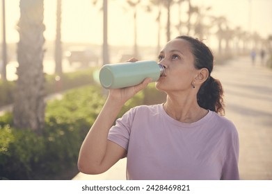 Curly haired Latina woman in purple t-shirt, drinking water from a steel bottle, standing on the treadmill and resting after jog at sunset. People. Active and healthy lifestyle. Sport. Cardio workout - Powered by Shutterstock