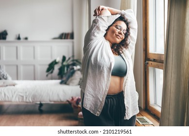 Curly Haired Overweight Young Woman In Blue Top And Shorts With Satisfaction On Face Accepts Curvy Body Shape In Stylish Bedroom. 