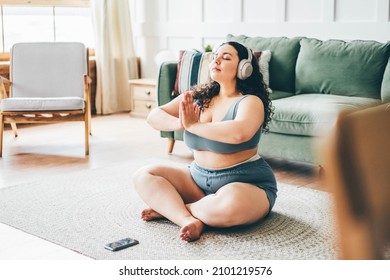 Curly haired overweight young woman in top and shorts meditating on floor mat against green sofa.