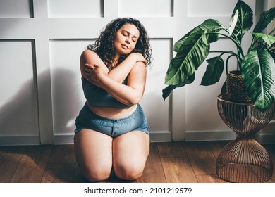 Curly haired overweight young woman in blue top and shorts with satisfaction on face accepts curvy body shape in stylish bedroom. 