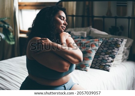 Curly haired overweight young woman in blue top and shorts with satisfaction on face accepts curvy body shape in stylish bedroom. 
