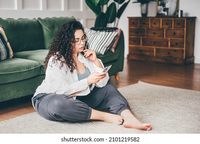 Curly haired plus size young woman wearing comfortable clothes in glasses clicks on smartphone screen sitting on floor mat in stylish room  - Powered by Shutterstock