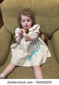 Curly Haired Toddler Sitting In Big Comfy Chair Eating Candy Lollipop With Spring Flower Dress On