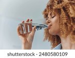 Curly haired woman drinking water from a glass with a refreshing look on her face