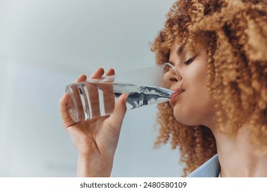 Curly haired woman drinking water from a glass with a refreshing look on her face - Powered by Shutterstock