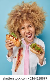 Curly Haired Woman Enjoys Eating Cheat Meal Holds Sandwich And Hot Dog Enjoys Favorite Fast Food Exclaims Loudly Wears White Shirt And Tie Stands Dirty Being Careless Very Hungry. Unhealthy Dieting