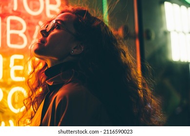 Kurz haarige junge Touristinnen mit leichtem Make-up in Gläsern sieht sich um Stehen in der Nähe von Bar mit buntem Neonzeichen gegen Nachtmegalopolis