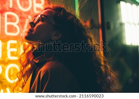 Curly haired young woman tourist with light makeup in glasses looks around standing near bar with colorful neon sign against night megalopolis