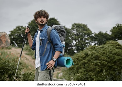 curly indian backpacker holding trekking poles on path in woods, hiker having adventure concept