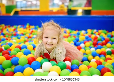 Curly Little Girl Having Fun In Ball Pit With Colorful Balls. Child Playing On Indoor Playground. Kid Jumping In Ball Pool.