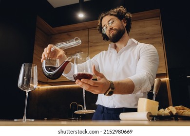 Curly man at home in a beautiful kitchen pours red wine into a glass with decanters. A sommelier pouring red wine into decanter