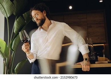 Curly man at home in a good kitchen holding a glass of red wine and looking at him. On the background of a beautiful strelitzia flower - Powered by Shutterstock