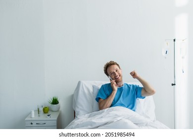 Curly Man In Patient Gown Talking On Smartphone In Hospital