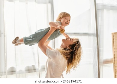 A curly mother joyfully lifts her toddler daughter up into the air, expressing love and playfulness at home. - Powered by Shutterstock