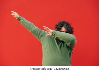 Curly Overweight Young Man Showing Dab Gesture On Red Background. Fat Guy In Green Sweatshirt Dancing Dab Dance On Red Wall Background.