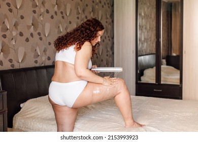 Curly Plus Size Woman In White Underwear Stand Near Bed In Bedroom And Wrap Hip With Cling Film Back View. Fat Burning Treatment Of Thick Thighs And Legs. Weight Loss Program, Body Wrap Procedure.