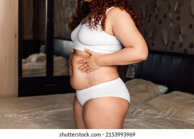 Curly plus size woman in white underwear standing near bed in bedroom and touching stomach with stretch marks closeup side view. Fat burning treatment of thick belly. Weight loss program, dieting.