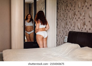 Curly plus size woman in white underwear look in wardrobe mirror near bed in bedroom and touch sagging thick stomach back view. Fat burning treatment of overweight body. Weight loss and dieting.