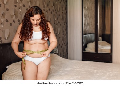 Curly plus size woman in white underwear stand near bed in bedroom, measure waist circumference with tape and surprise. Overweight girls struggle with obesity. Weight loss program, body control.