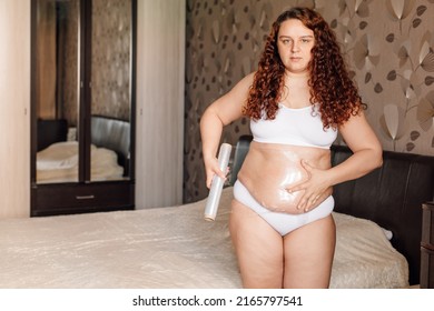 Curly Plus Size Young Woman In White Underwear Stand Near Bed In Bedroom And Wrap Sagging Belly With Cling Film. Fat Burning Treatment Of Thick Stomach. Weight Loss Program, Body Wrap Procedure.