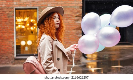 Curly Red-haired Girl Wear Casual Felt Hat and Cloak Raincoat Walking on City Street. Pretty Young Woman Holds Many Air Balloons in Spring Cloudy Evening. Rainy Mood