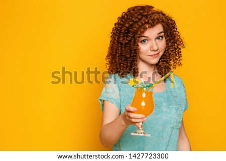 curly red-haired woman with a glass of summer cocktail on a yellow background.