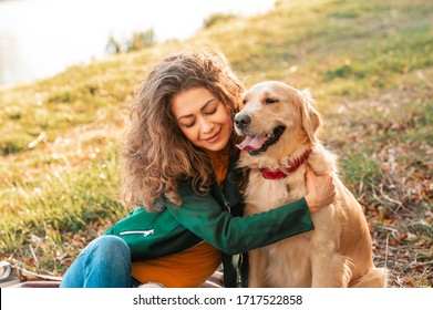 Curly Smiling Girl Embrance Her Pet Dog Near Face. Golden Retriever Dog Playing With A Curly Woman Walking Outdoors Sunny Day. Love And Care For The Pet.
