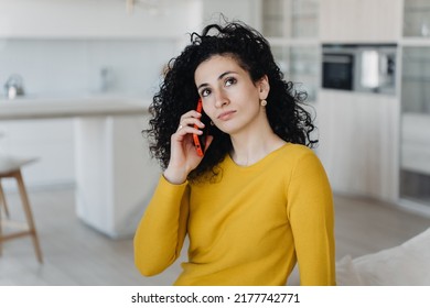 Curly Spanish Young Woman In Yellow Sweater Talking By Phone Remote Working At Home On Cozy Sofa. Purposeful Hispanic Female Entrepreneur Speaking With Business Partner.  Caucasian Housewife At Home.