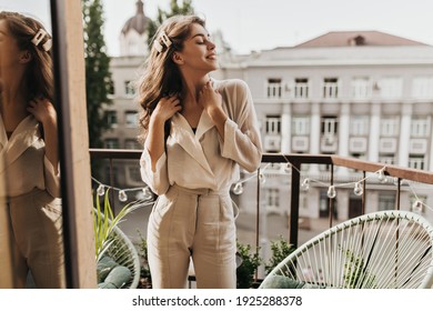 Curly woman enjoys sunny day on terrace. Pretty dark-haired lady in beige blouse and stylish pants poses on balcony - Shutterstock ID 1925288378