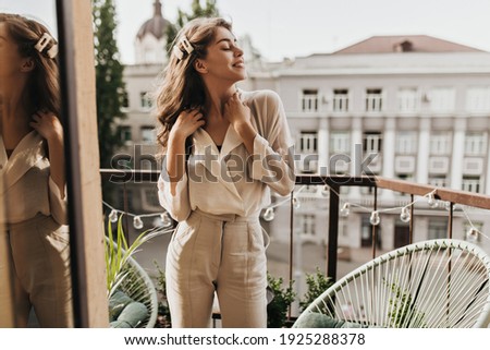 Curly woman enjoys sunny day on terrace. Pretty dark-haired lady in beige blouse and stylish pants poses on balcony