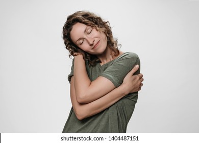 Curly woman hugging herself, looks happy, loves herself, has high self esteem, smiles from pleasure. Lovely calm peaceful cheery girl isolated over white. Love your self and self acceptance concept.