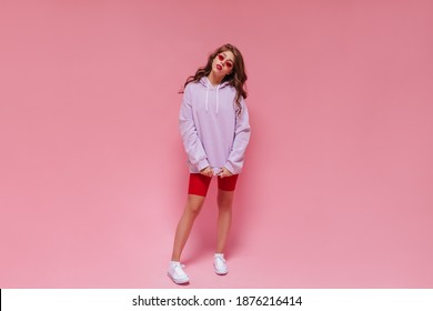 Curly Woman In Red Cycling Shorts And Purple Hoodie Looks Into Camera. Charming Girl In Sunglasses Poses On Pink Background.