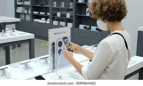 Curly Woman In White Face Mask Protecting From Coronavirus On Electronic Shopping, Making Selfie On A New IPhone 13 In A Store, With IPhone 13 Pro Sierra Blue Plate In The Background 