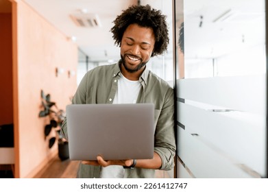 Curly young african-american businessman standing in office hallway and using laptop for office work, male employee checks mail, conducts business correspondence with customers - Shutterstock ID 2261412747