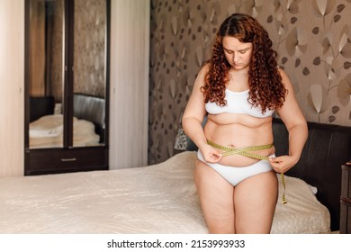 Curly young plus size woman in white underwear stand near bed in bedroom and measure waist circumference with tape. Overweight girls struggle with obesity. Weight loss program, body control.